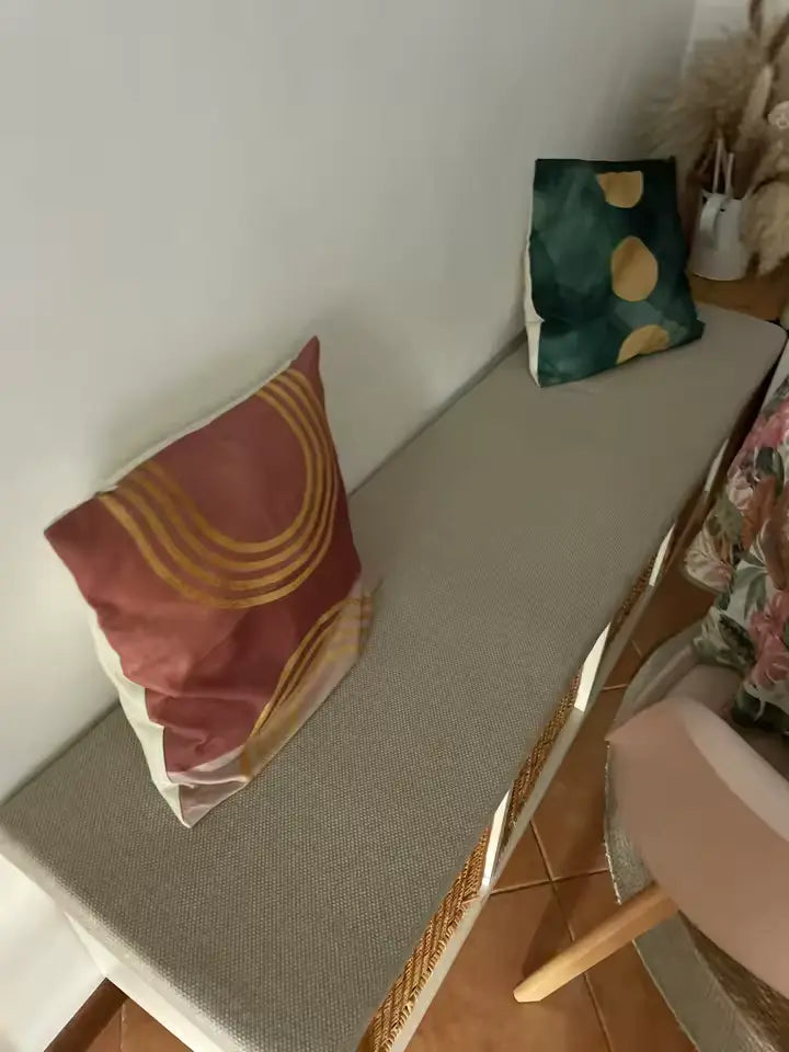 Un banc moelleux rembourré gris texturé avec un cadre en osier tissé et deux coussins décoratifs jetés—un en terre cuite chaude avec des rayures courbes dorées et un autre en vert profond avec des pois moutarde-jaune.